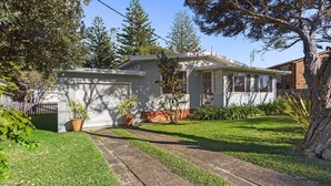 A single-story house with a garage, a front yard with a lawn, and a large tree to the right. - Between 2 Beaches – pet friendly cottage (Culburra Beach)