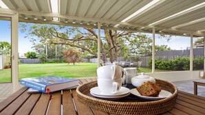 A covered patio area with a table set for tea, including a book, teacups, a teapot, sugar bowl, and a slice of cake on a tray. - Sea Bird – unplug unwind and enjoy (Culburra Beach)