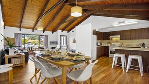 A spacious dining area with a wooden table and chairs, a kitchen with dark wooden cabinets, and a view of the outdoors. - Sea Bird – unplug unwind and enjoy (Culburra Beach)