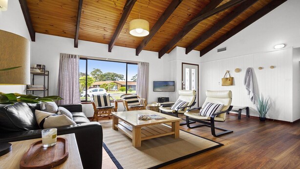 A spacious living room with a wooden ceiling, leather sofa, and a large window offering a view of the outside. - Sea Bird – unplug unwind and enjoy (Culburra Beach)