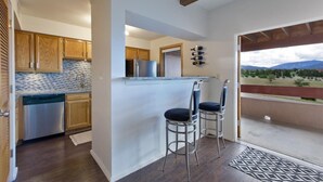 Una cocina moderna con armarios de madera, lavavajillas de acero inoxidable y salpicadero de azulejos. Hay dos taburetes de bar y se ve una vista de las montañas a través de una puerta balcón.