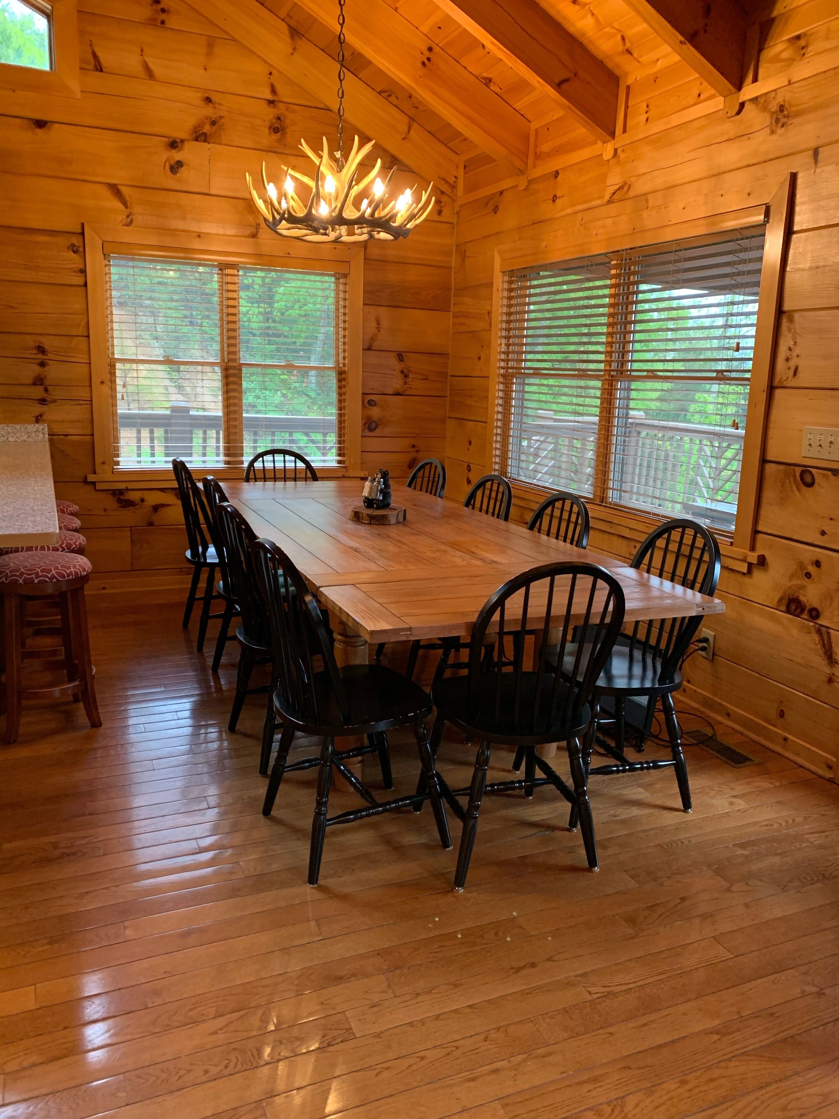 Une salle à manger en bois, avec une grande table et des chaises, un lustre et des fenêtres équipées de stores.