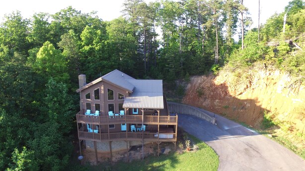A large wooden cabin with multiple balconies, surrounded by lush greenery and a winding road. - Breathtaking mountain and sunset views...huge cabin with cozy atmosphere (Pigeon Forge)