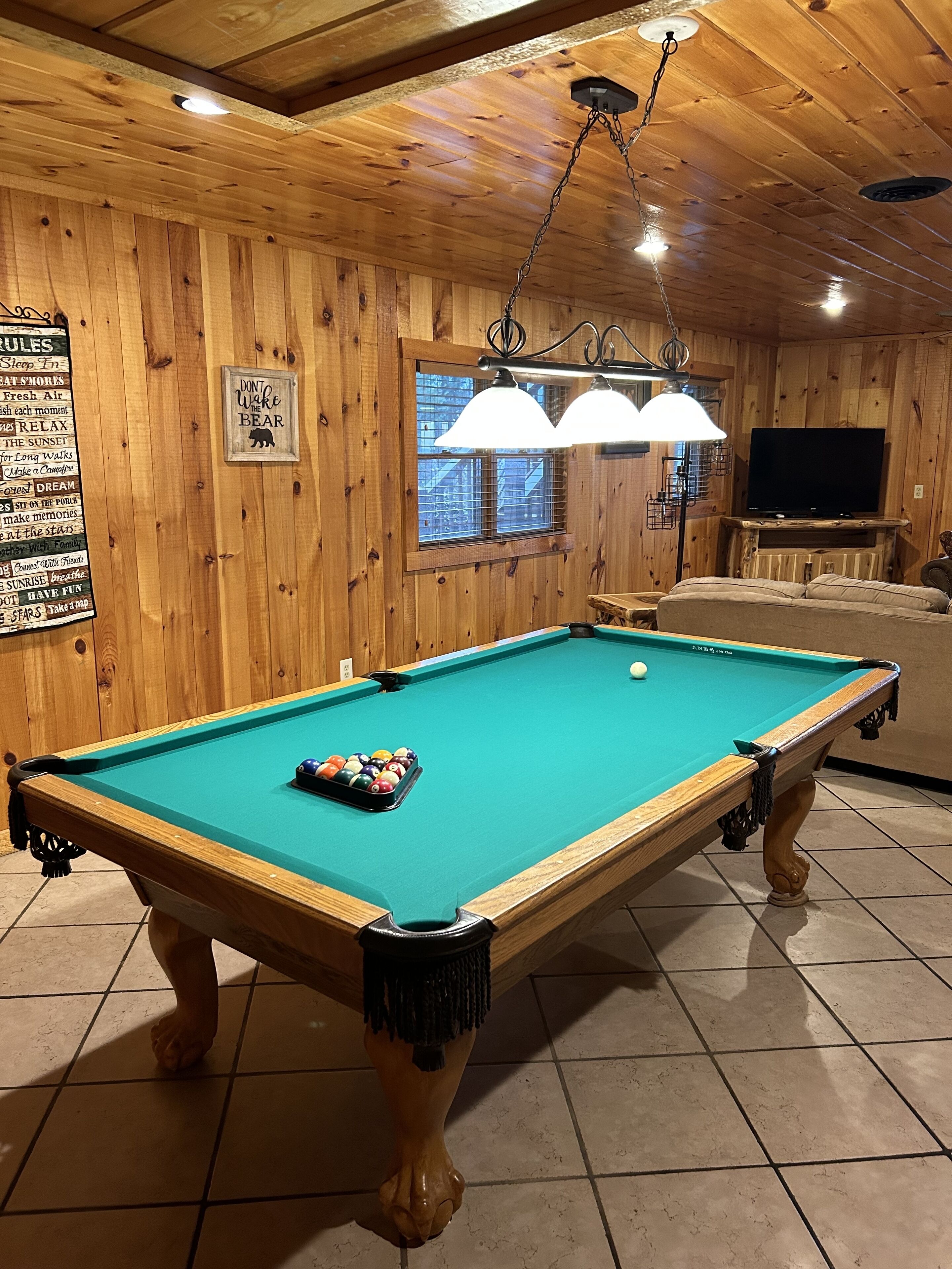 A pool table with a cue ball and a rack of billiard balls, a television mounted on the wall, and a wooden ceiling with a hanging light fixture.