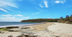 Una playa con un primer plano rocoso, una orilla arenosa y una colina boscosa al fondo.