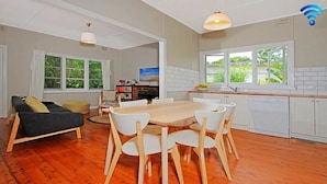 A modern kitchen with white cabinets, a wooden dining table, and chairs. There is a sofa and a television in the living area. - Culburra Sands – cosy cottage (Culburra Beach)