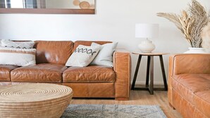 En brun lædersofa med puder, en rund vævet skammel, en lille hvid lampe på et træbord og en vase med tørrede planter.