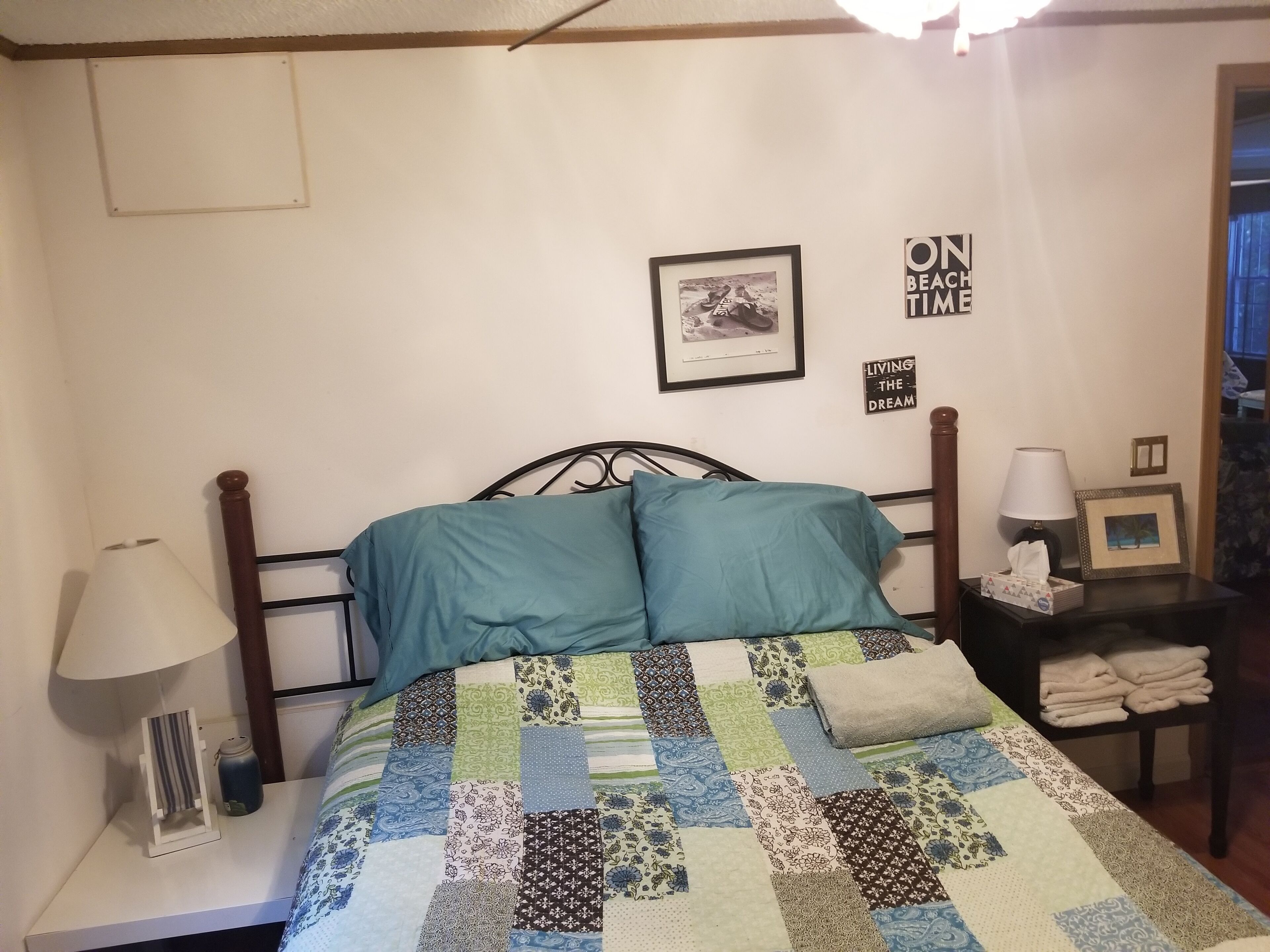 A bed with a patchwork quilt, a bedside lamp, a framed picture, and a small table with a tissue box and a lamp.