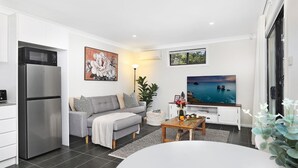 A modern living room with a grey sofa, a flat-screen TV, a kitchen area with a fridge, and a large window offering a view of the sea. - Ravello – Stay 3, Pay 2! (Callala Bay)