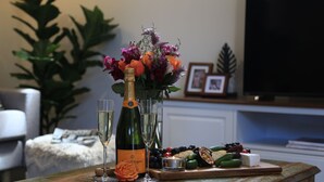 A table with a bottle of champagne, two glasses, a floral arrangement, and a cheese board with various items. - Ravello – Stay 3, Pay 2! (Callala Bay)