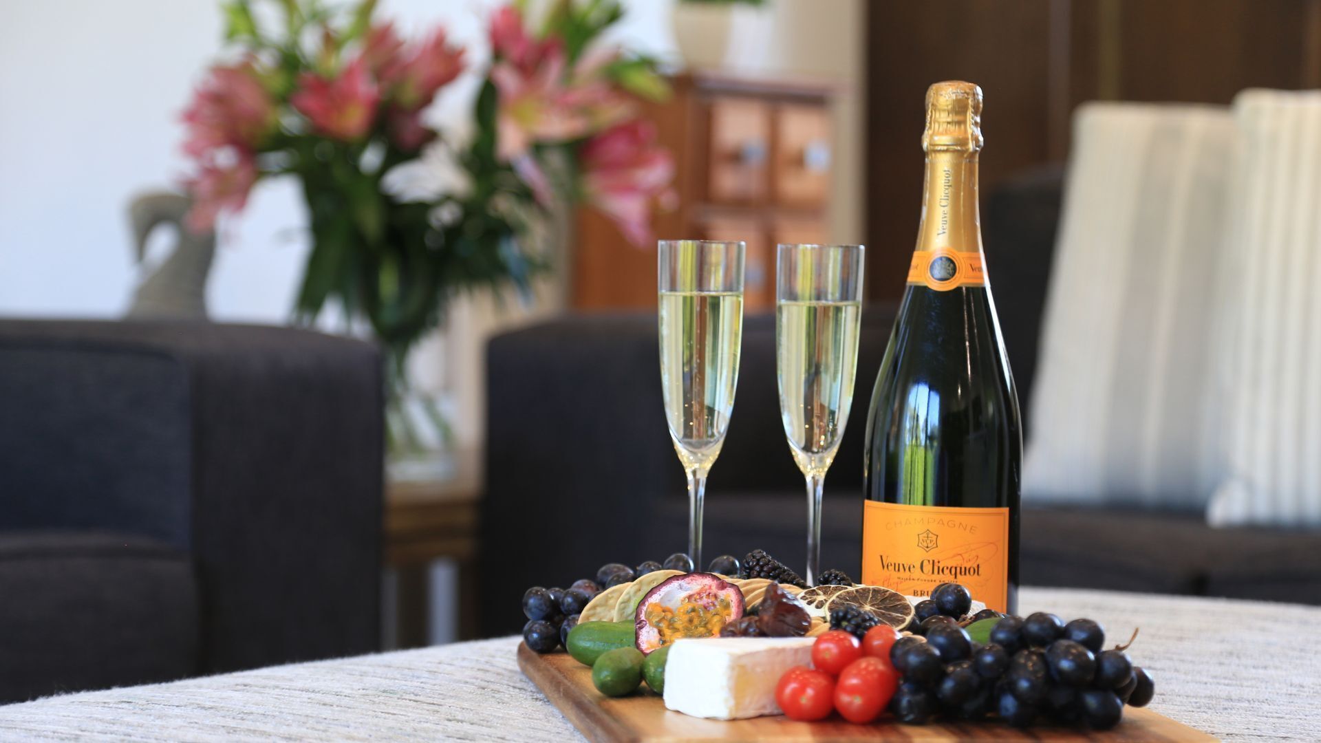 Pullo Veuve Clicquot -samppanjaa, kaksi lasia sekä puinen tarjoilulauta, jossa on juustoa, viinirypäleitä ja passionhedelmää.