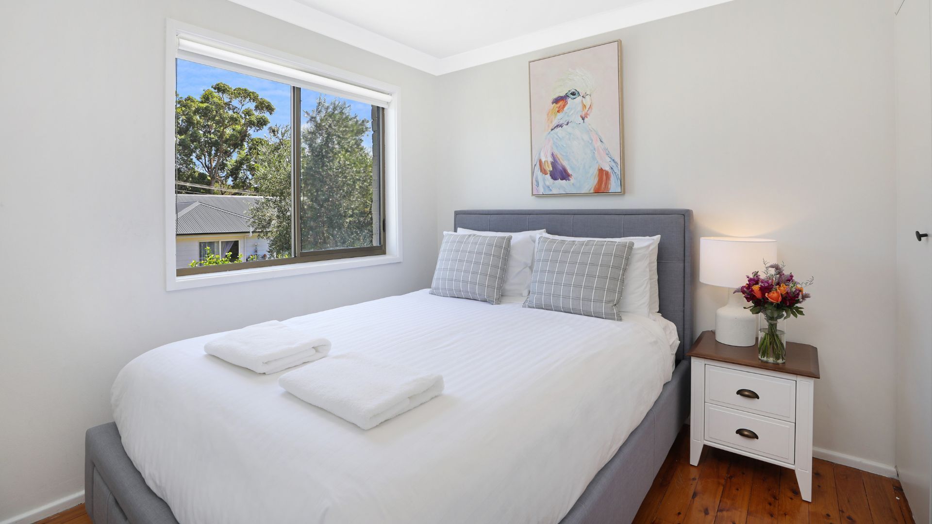 A neatly made bed with white linens and two pillows, a bedside table with a lamp and a vase of flowers, and a window with a view of trees and a house.