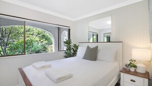 A modern bedroom with a large bed, a bedside table with a lamp, a mirror, and a view of greenery through a window. - Ravello – Stay 3, Pay 2! (Callala Bay)