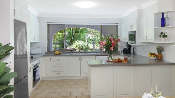 A modern kitchen with white cabinetry, a large island, a window with a view of greenery, and a wine glass on the counter. - Ravello – Stay 3, Pay 2! (Callala Bay)