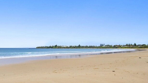 A wide sandy beach with gentle waves, a clear sky, and a distant shoreline with buildings and greenery. - Cleveland Place - beach access (Culburra Beach)
