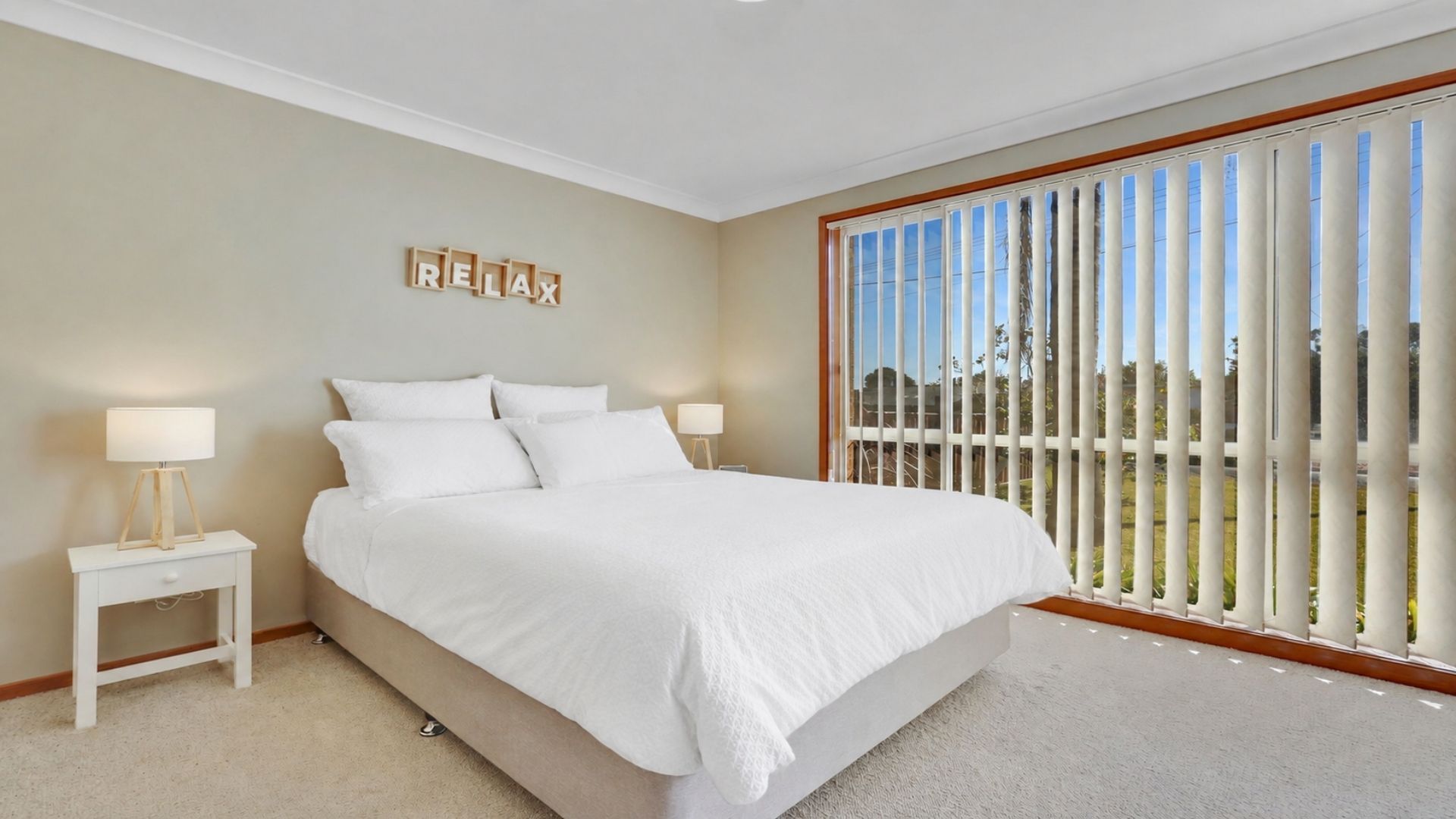 A bedroom with a large bed, two bedside tables with lamps, a view of the outdoors through a sliding door, and a 'RELAX' sign on the wall.