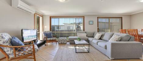 A living room with a sofa, a television, a coffee table, and a chair. There is a window with blinds and a view of the outside.