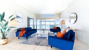 A modern living room with two blue sofas, a coffee table, and a large window offering a view of the sea.