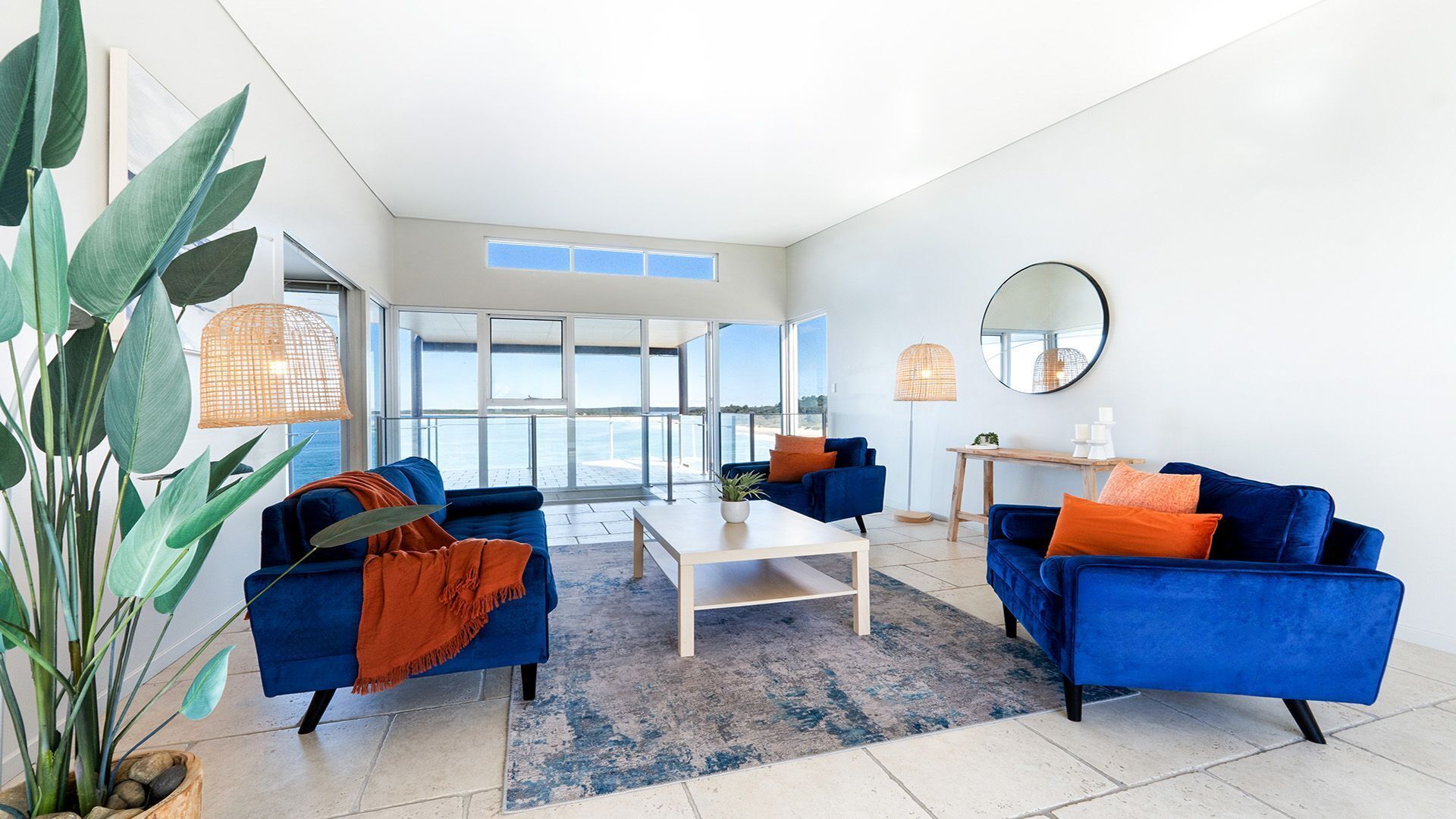 A modern living room with two blue sofas, a coffee table, and a large window offering a view of the sea.