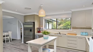 A modern kitchen with a built-in refrigerator, a window with a view of greenery, a dining area with a table and chairs, and a hanging light fixture. - Gone Fishing! - ideal for larger families and groups with pets (Culburra Beach)
