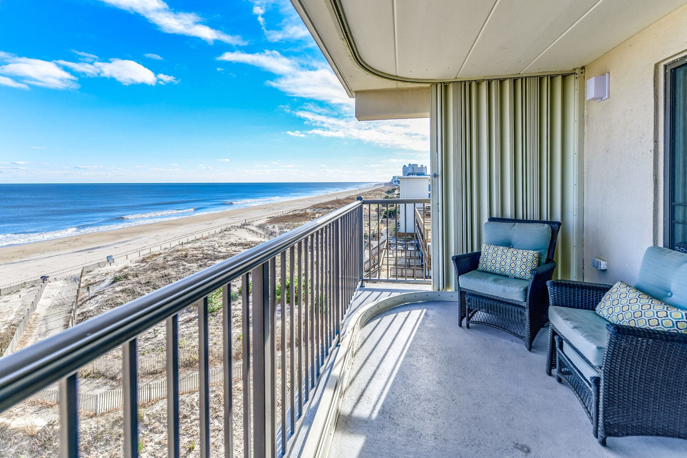 A balcony with wicker furniture, a view of the beach, and a building in the background.