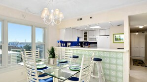 Een moderne keuken met witte kasten, een glazen eiland en een eethoek met witte stoelen en blauwe kussens.