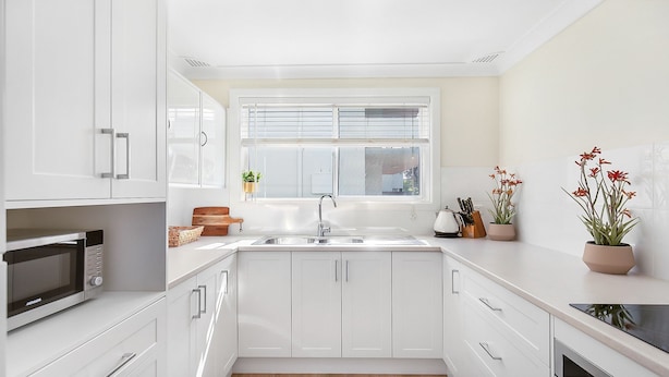 Een moderne keuken met witte kasten, een ingebouwde magnetron, een spoelbak met raam erboven en een potplant op het aanrecht.