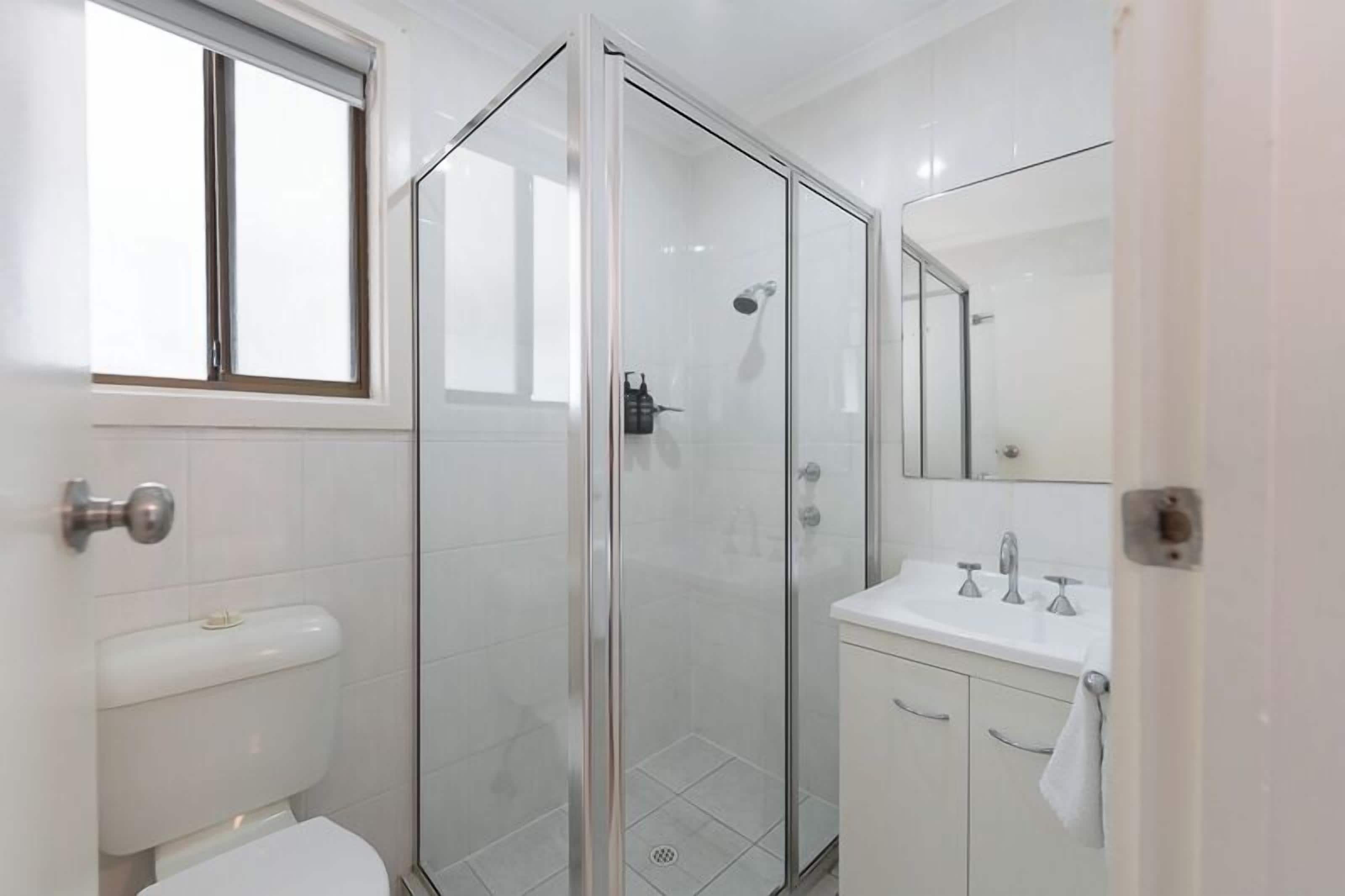 A modern bathroom with a glass-enclosed shower, a toilet, a sink with a mirror, and a window above the toilet.