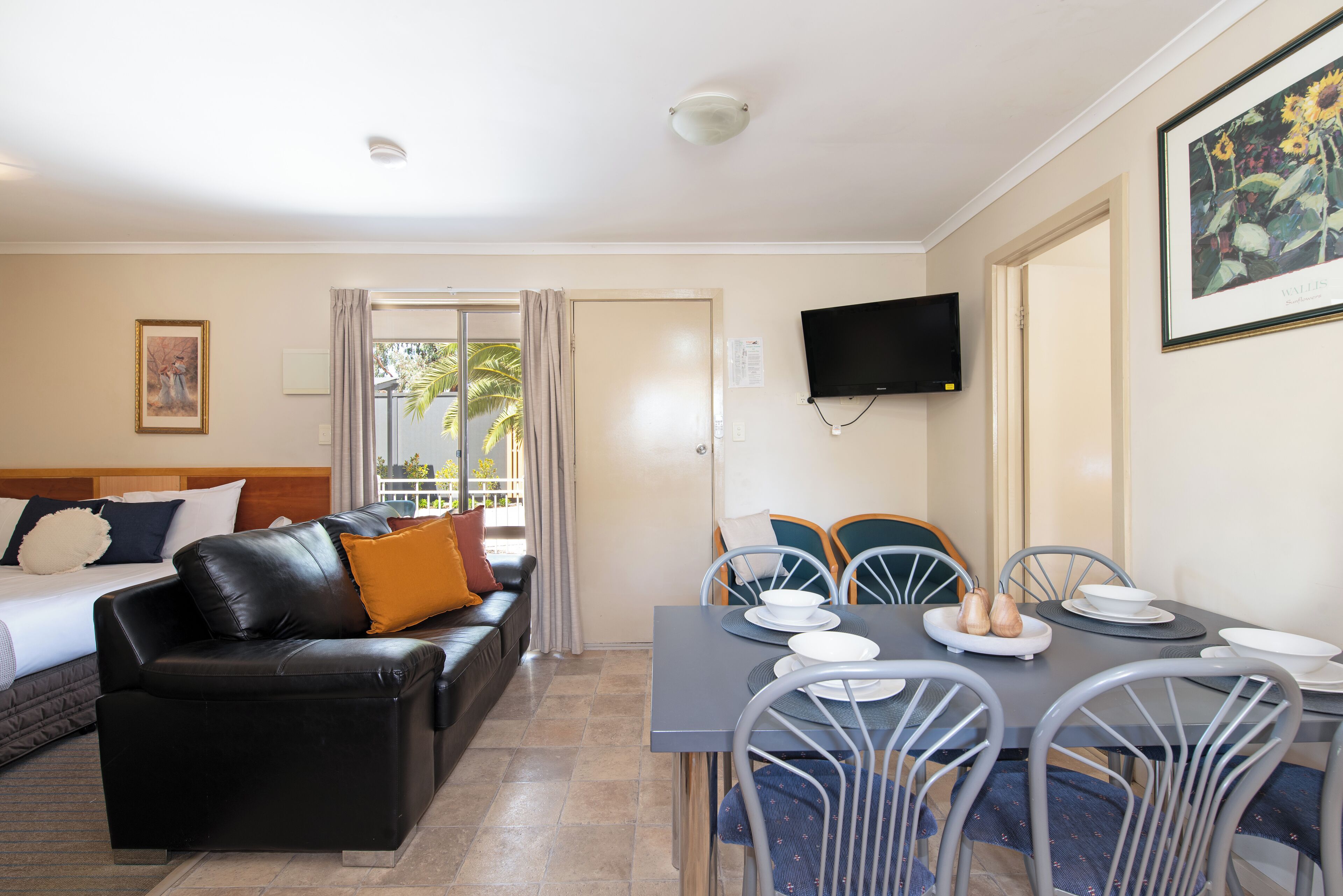 A hotel room with a bed, a black leather sofa, a dining table with chairs, and a TV mounted on the wall.