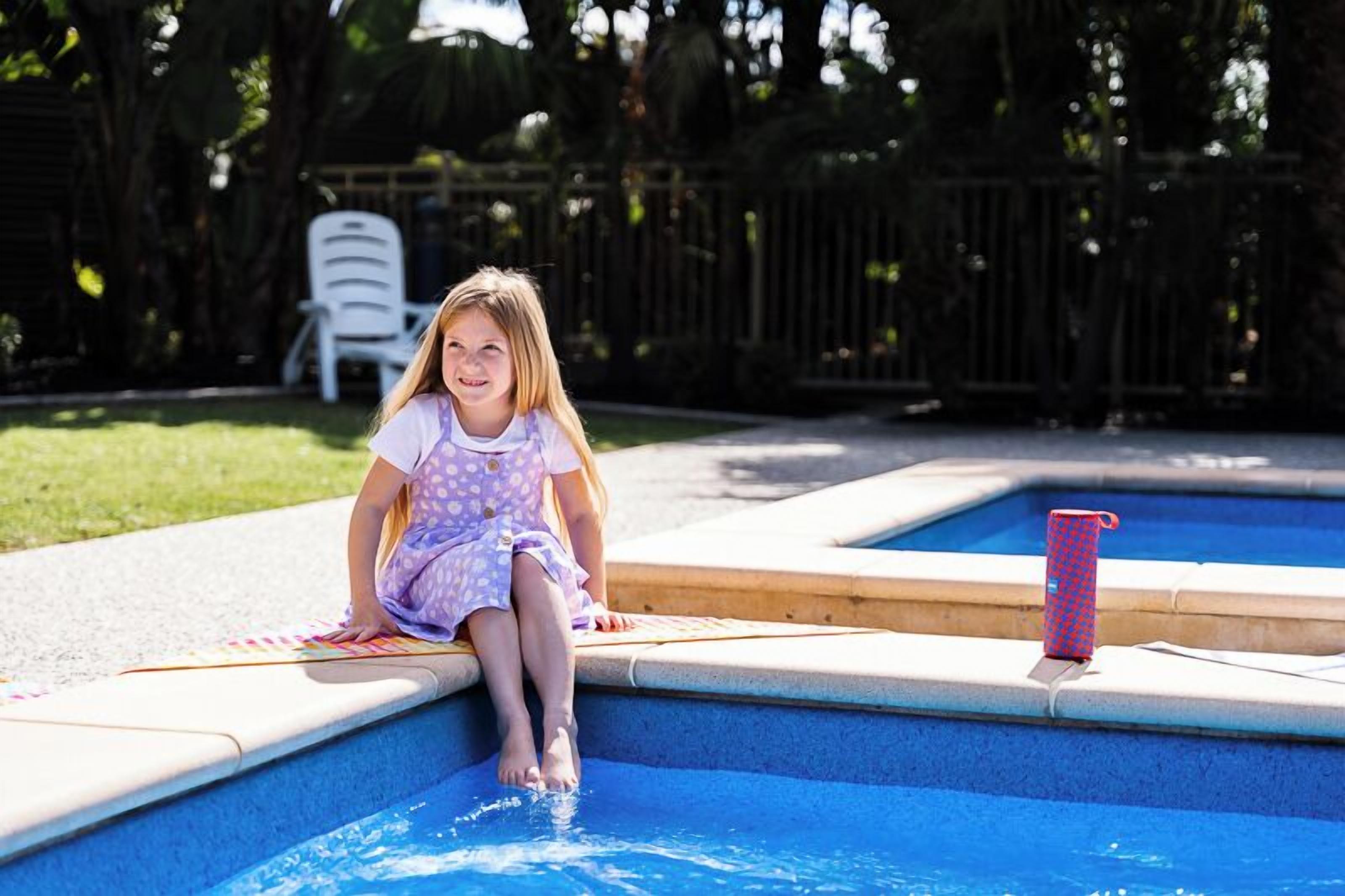 Un enfant est assis au bord d’une piscine, avec une chaise et un objet rouge et bleu en arrière-plan.