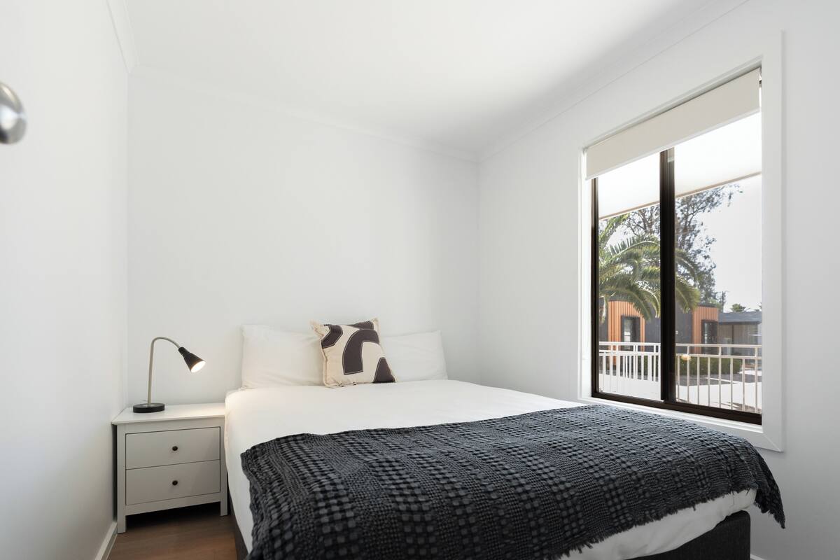 A bedroom with a bed, a nightstand, a lamp, and a window with a view of palm trees.