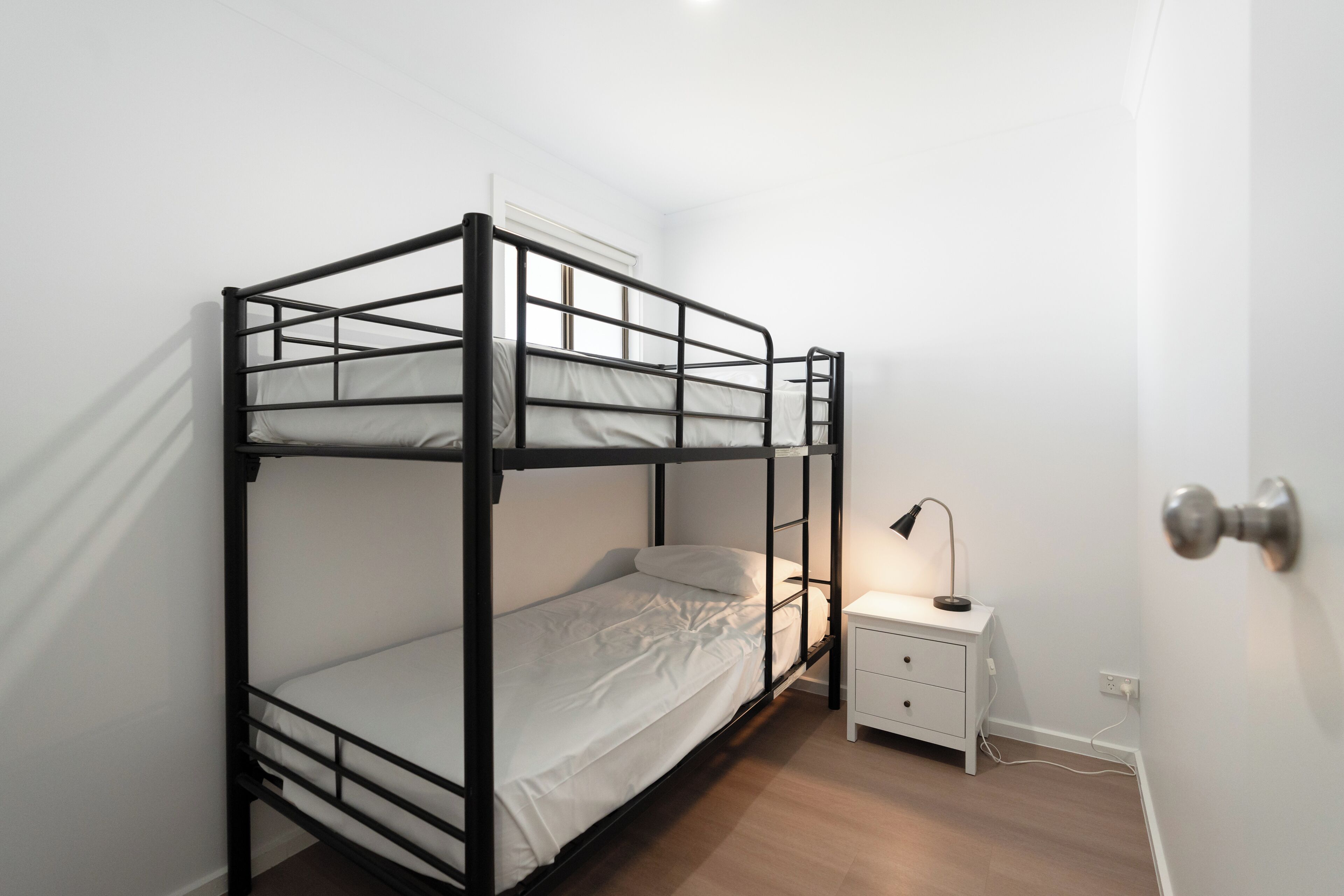 Habitación con literas, una pared blanca, una mesita de noche con lámpara y una puerta con pomo plateado.