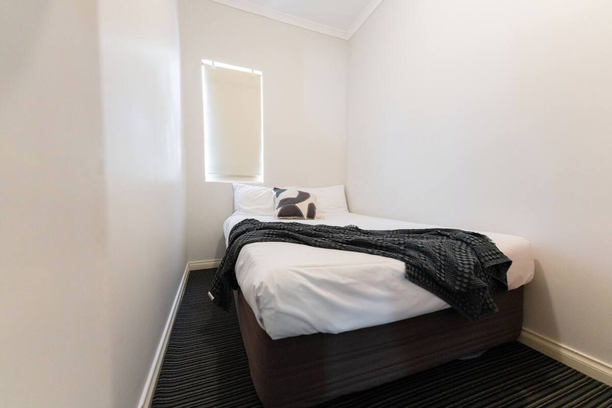 A small, neatly arranged bedroom with a single bed, a window, and a carpeted floor.