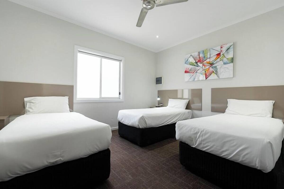 A hotel room with three single beds, a ceiling fan, a large abstract painting, and a window providing natural light.