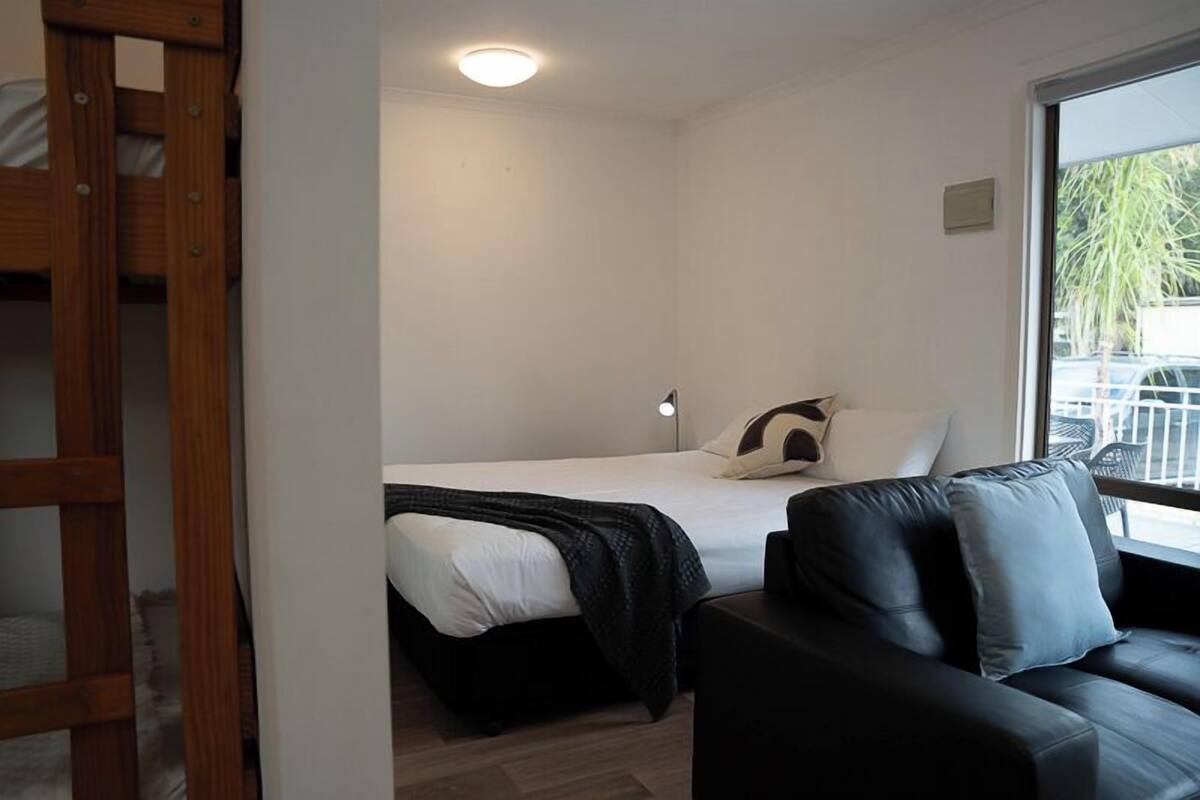 A bedroom with a bed, a black leather sofa, a wooden ladder, and a window with a view of parked cars.