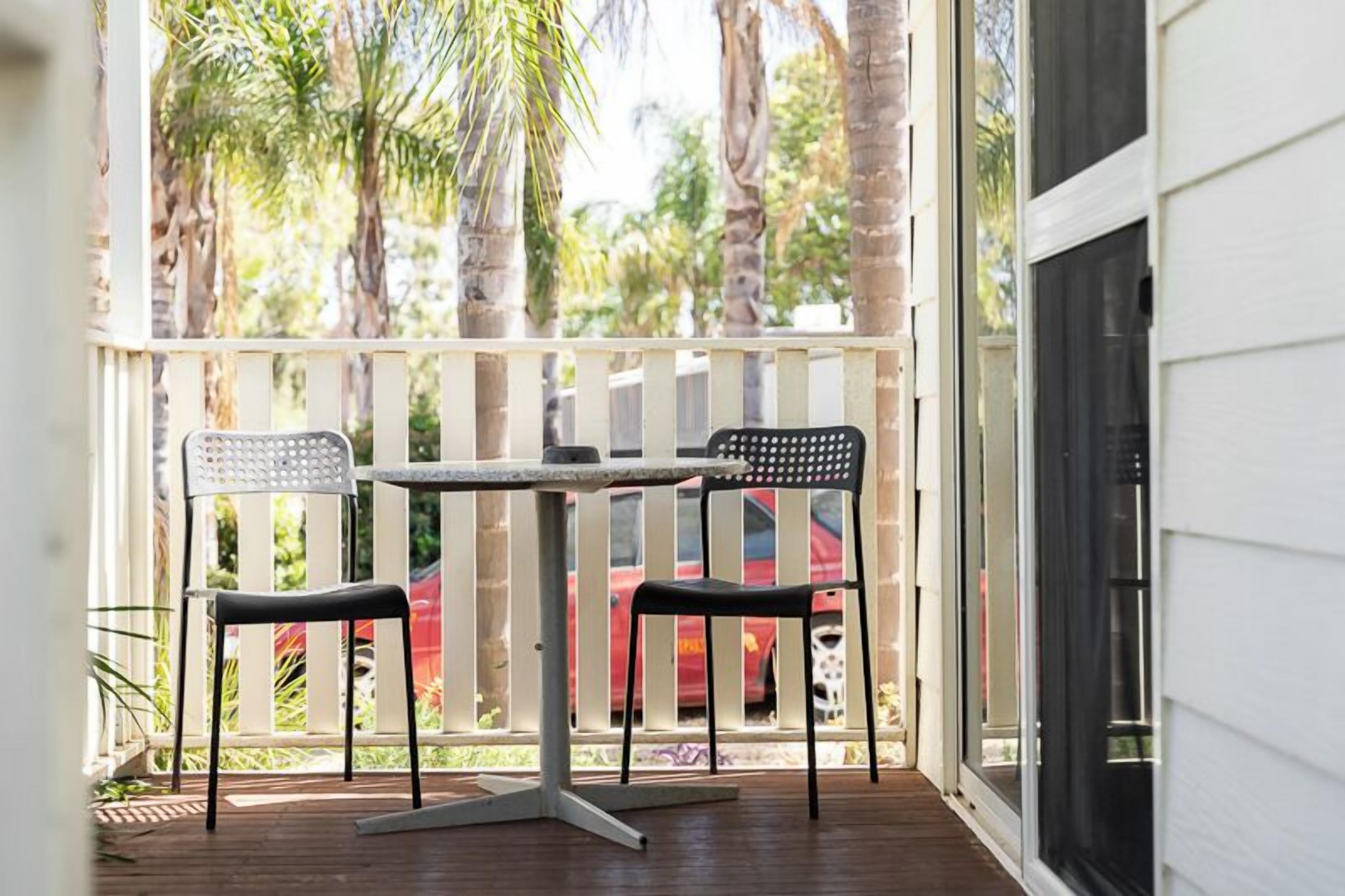 A covered outdoor area with a table and two chairs, a white railing, and a view of palm trees and a red car in the background.