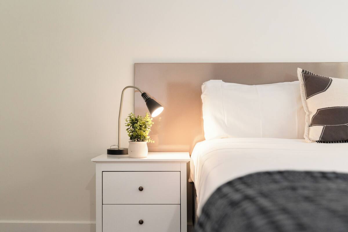A neatly made bed with white bedding and a black throw blanket, a bedside lamp, and a small potted plant on a white nightstand.