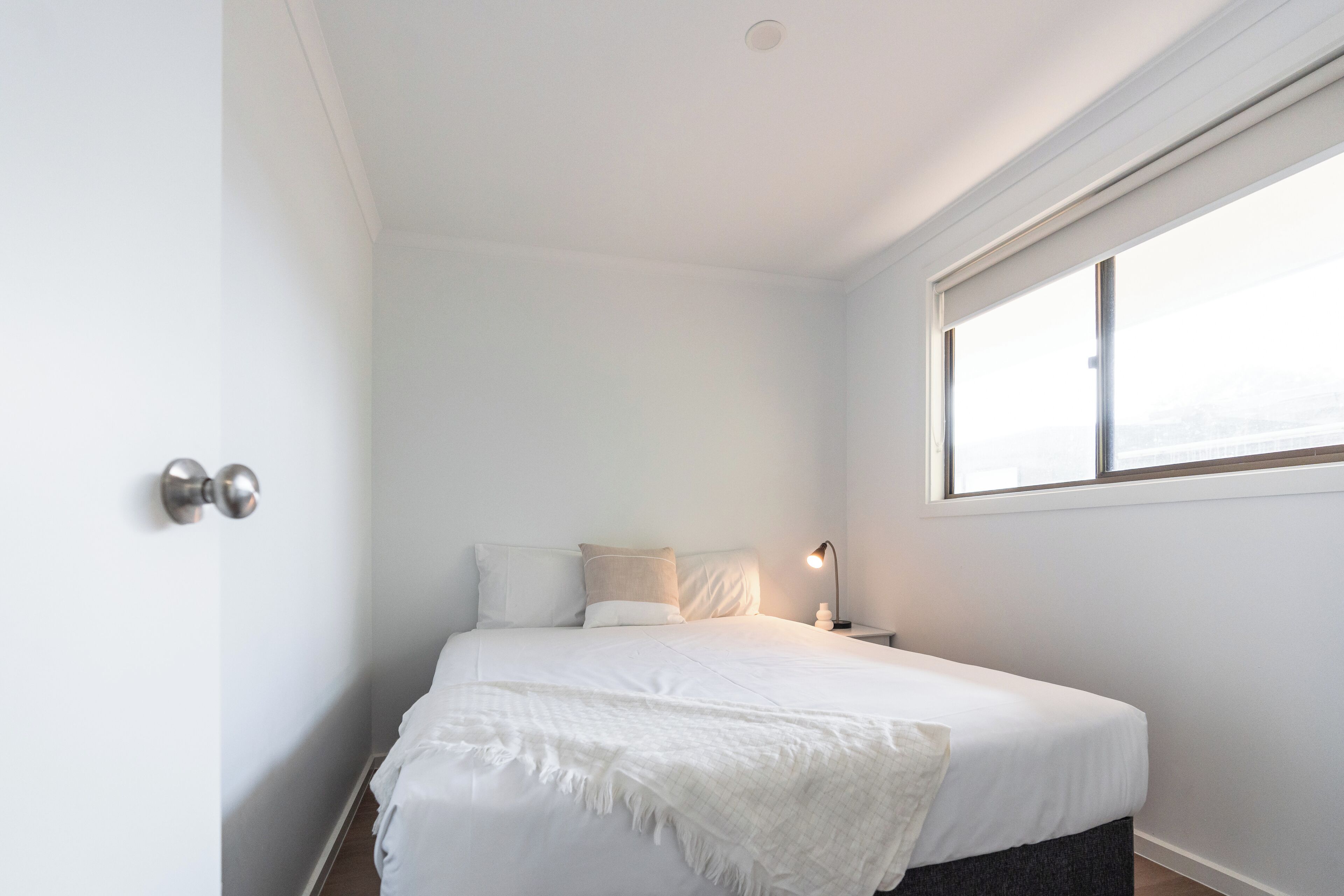 Een minimalistische slaapkamer met een groot bed, een bedlampje en een raam dat natuurlijk licht binnenlaat.