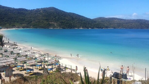 Ein Strand mit klarem türkisblauem Wasser, einem Sandufer und einer Reihe von Strandsonnenschirmen.