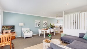 A modern living room with a dining area, a grey sofa, a wooden coffee table, and a TV on a wooden stand. - Ocean Blue Apartment Three (Currarong)