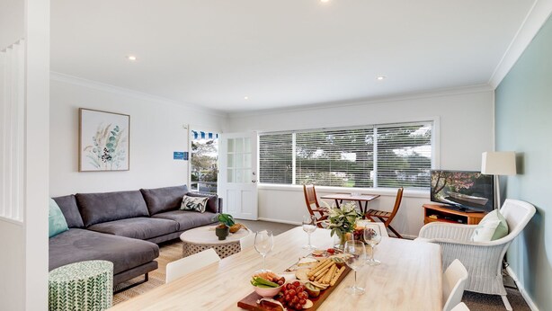 A spacious living room with a grey sofa, a dining table set with food and wine glasses, and a TV on a wooden stand. - Ocean Blue Apartment Three (Currarong)