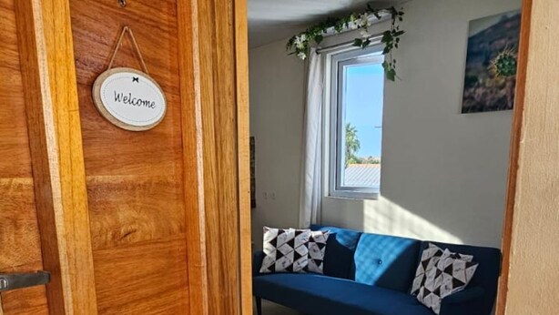 Une porte en bois ornée d’une pancarte « Bienvenue », qui donne sur une pièce avec un canapé bleu et des coussins à motifs, une fenêtre avec des rideaux et un paillasson « Bienvenue » à l’entrée.