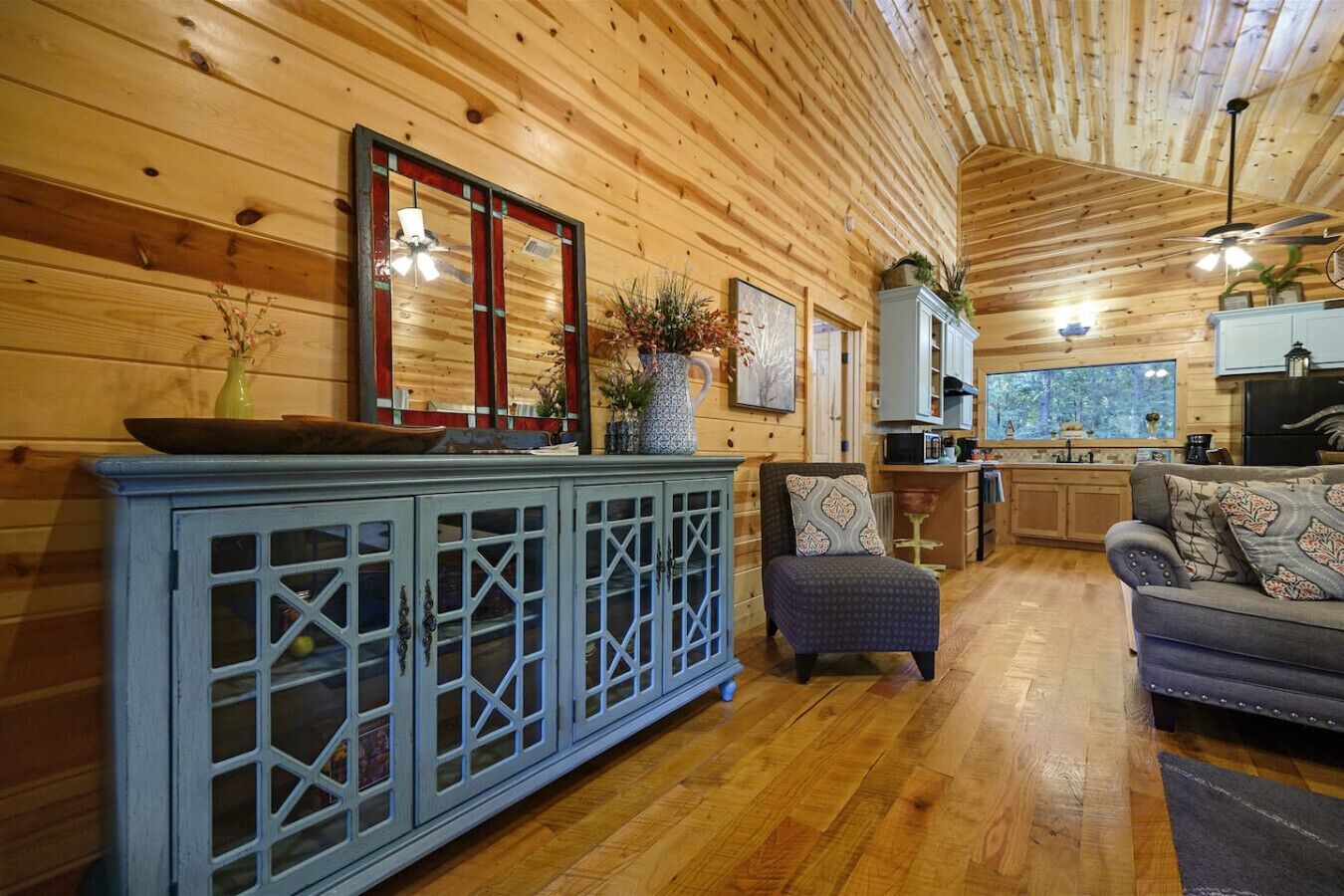 A cozy cabin interior with a wooden ceiling, a dining area with a blue cabinet, a gray sofa, and a kitchen with a large window.