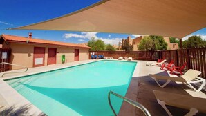 A swimming pool with lounge chairs and a shaded area, surrounded by a wooden fence and buildings. - Adobe Dreams - 3292, Private hot tub (Moab)