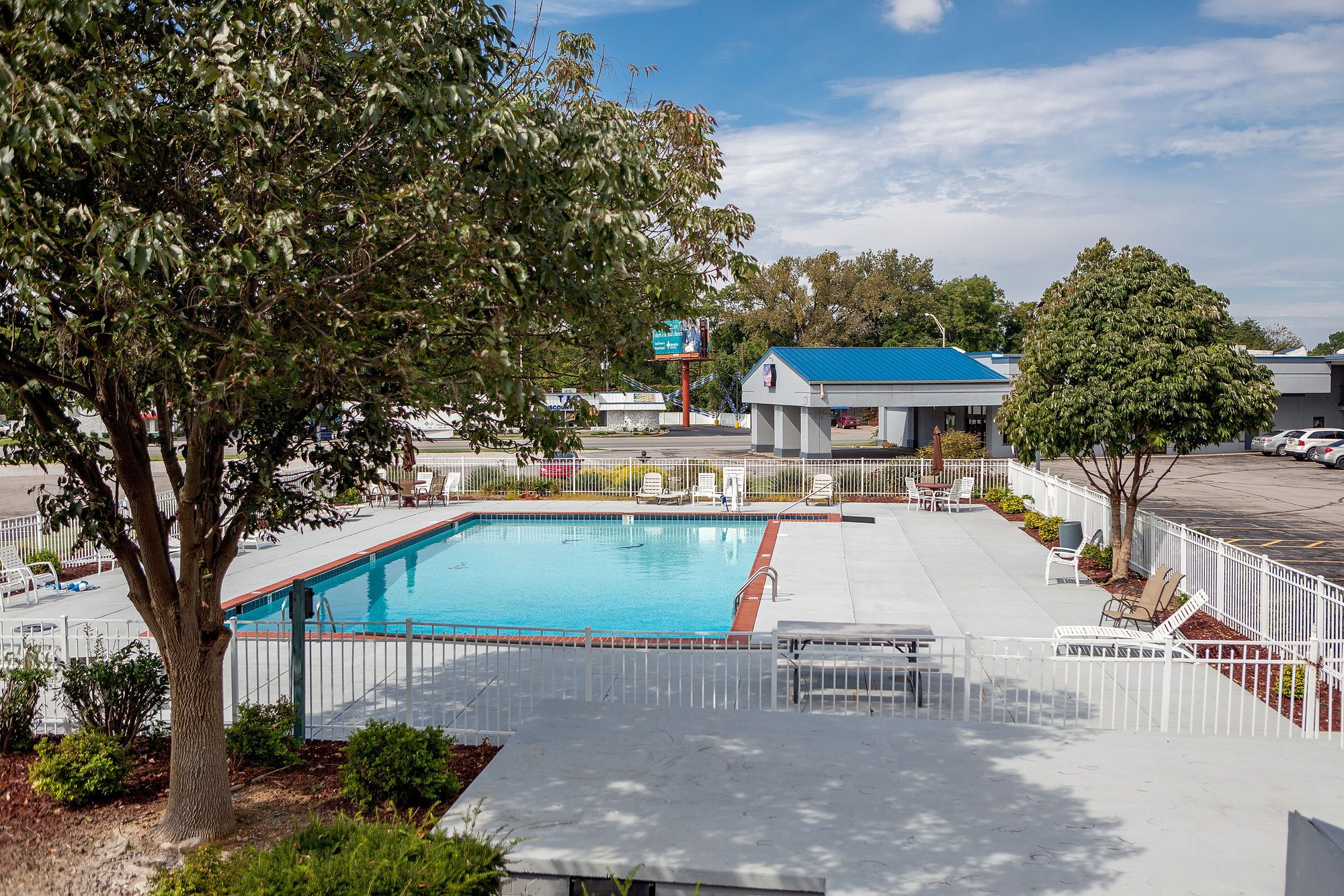 A swimming pool with a white fence, lounge chairs, and a building with a blue roof in the background.