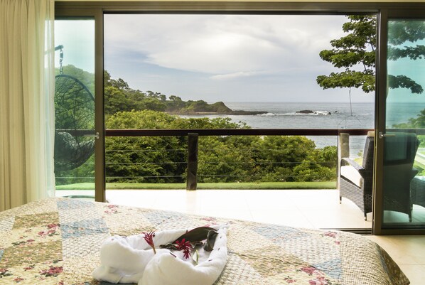 Ein Schlafzimmer mit großem Fenster und Blick auf eine Küstenlandschaft, einem Bett mit floralem Überwurf und einem Sessel am Fenster.