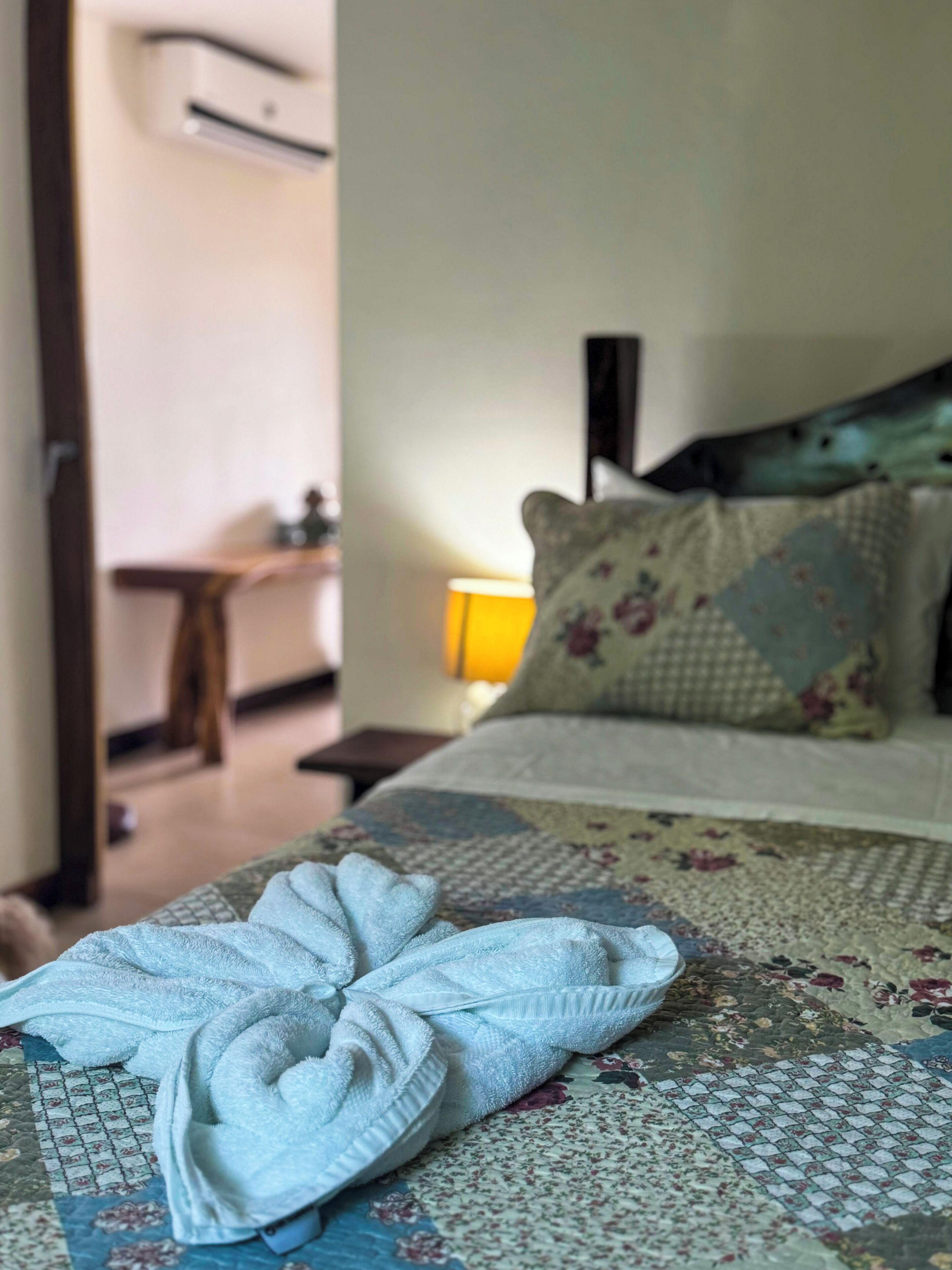 A neatly made bed with a floral-patterned bedspread, a bedside lamp, and a folded towel on the bed.