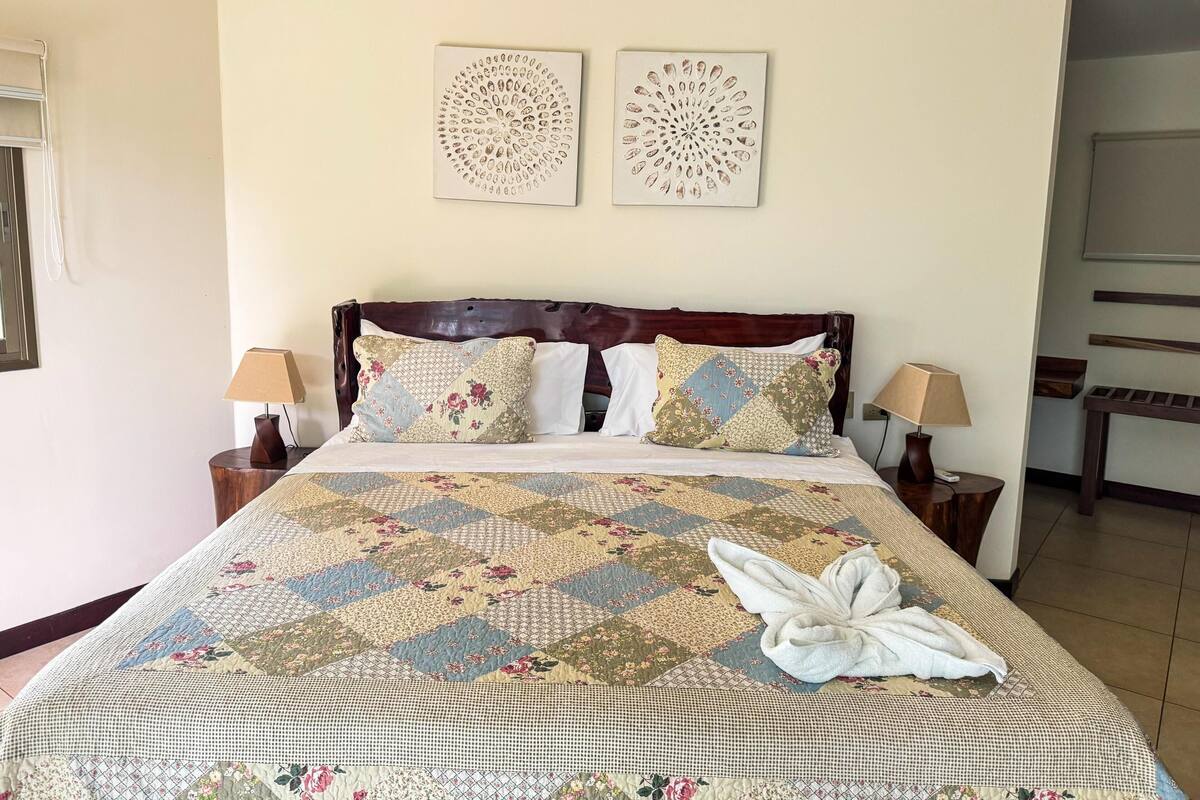Una cama bien hecha con un edredón estampado con motivos florales, dos almohadas decorativas y una toalla doblada. Hay dos lámparas de noche y una mesita de madera.