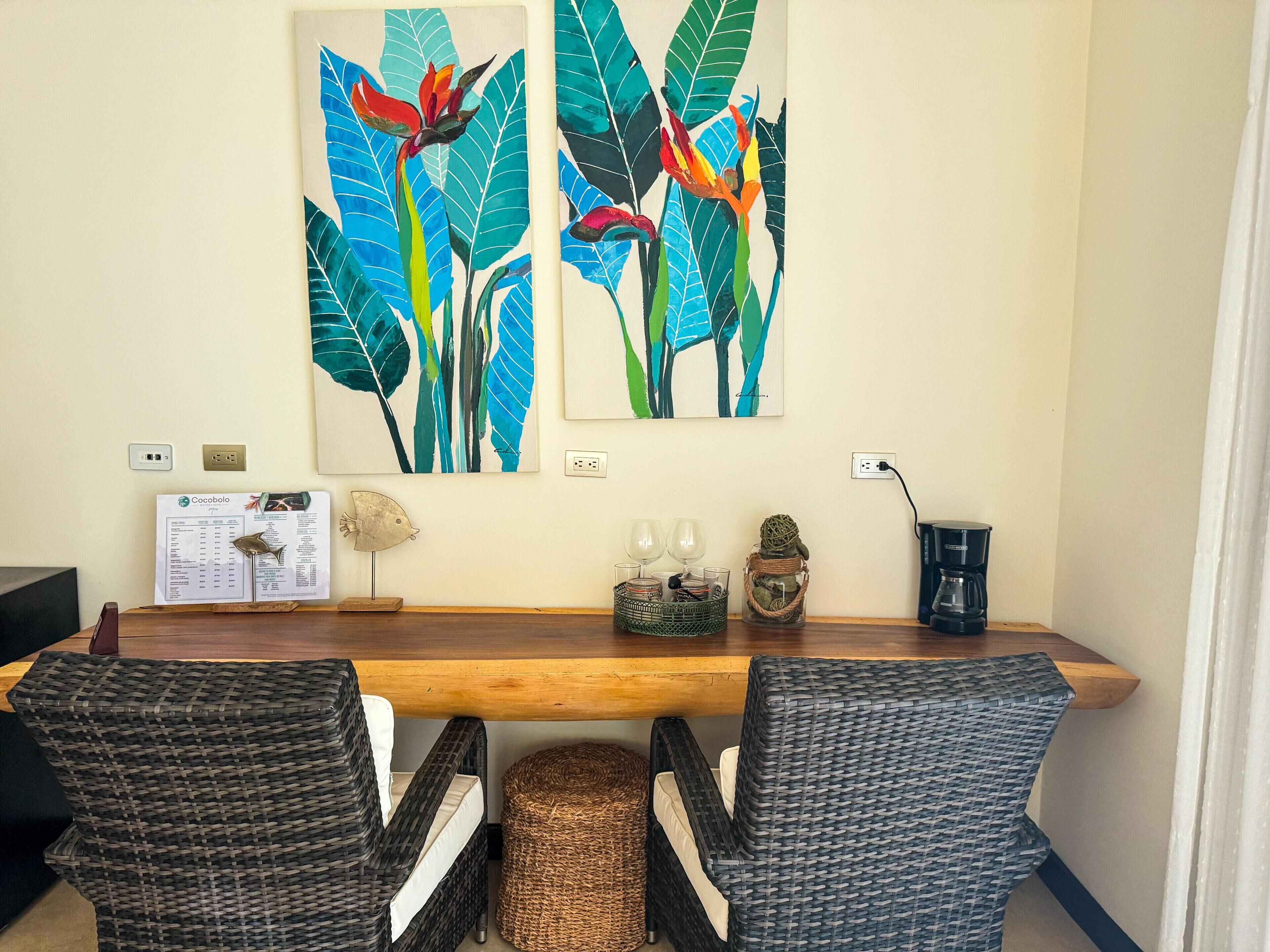 Ein Essbereich mit Holztisch, zwei Korbstühlen, einer Speisekarte, einer dekorativen Fischskulptur, einem gewebten Korb und zwei an der Wand befestigten Bildern von tropischen Blättern und Blumen.
