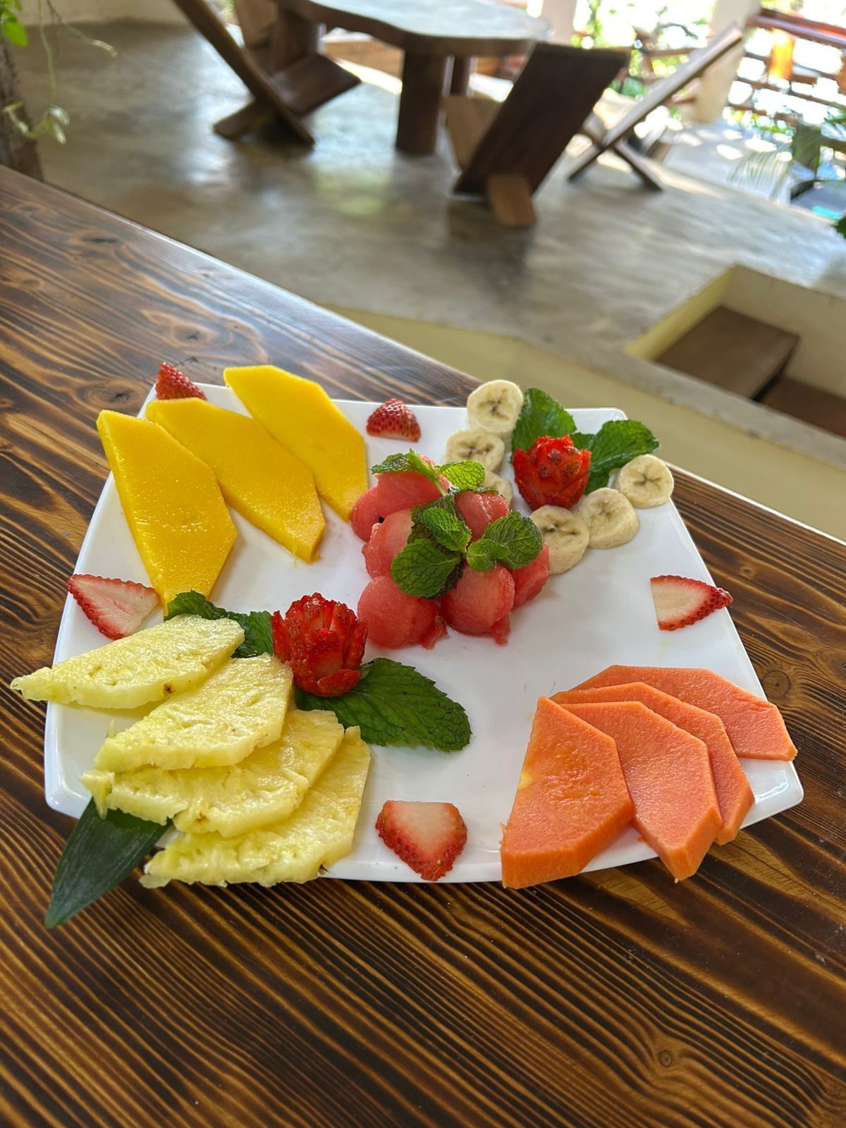 Eine Obstplatte auf einem weißen Teller mit Mangostreifen, Ananas, Erdbeeren, Banane und Wassermelone, garniert mit Minzblättern.